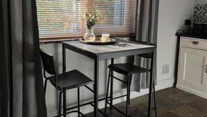 Eine kleine Küchenzeile mit einem schwarzen Metalltisch und zwei schwarzen Stühlen. Ein Fenster mit Jalousien, ein Blumenstrauß in einer Vase und eine Kerze auf dem Tisch sind zu sehen.