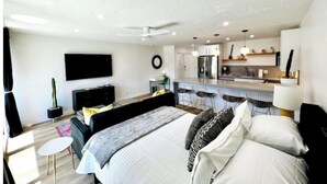 A modern bedroom with a large bed, a flat-screen TV, a desk, and a kitchenette in the background. - Location, Location, Location - Shadow Mountain Resort South Palm Desert  (Palm Desert)