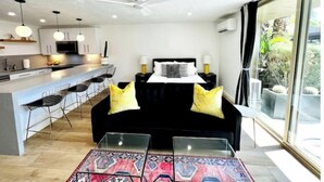A modern living area with a black sofa, a glass coffee table, and a patterned rug. There is a kitchen area with white cabinets and a bar counter. - Location, Location, Location - Shadow Mountain Resort South Palm Desert  (Palm Desert)