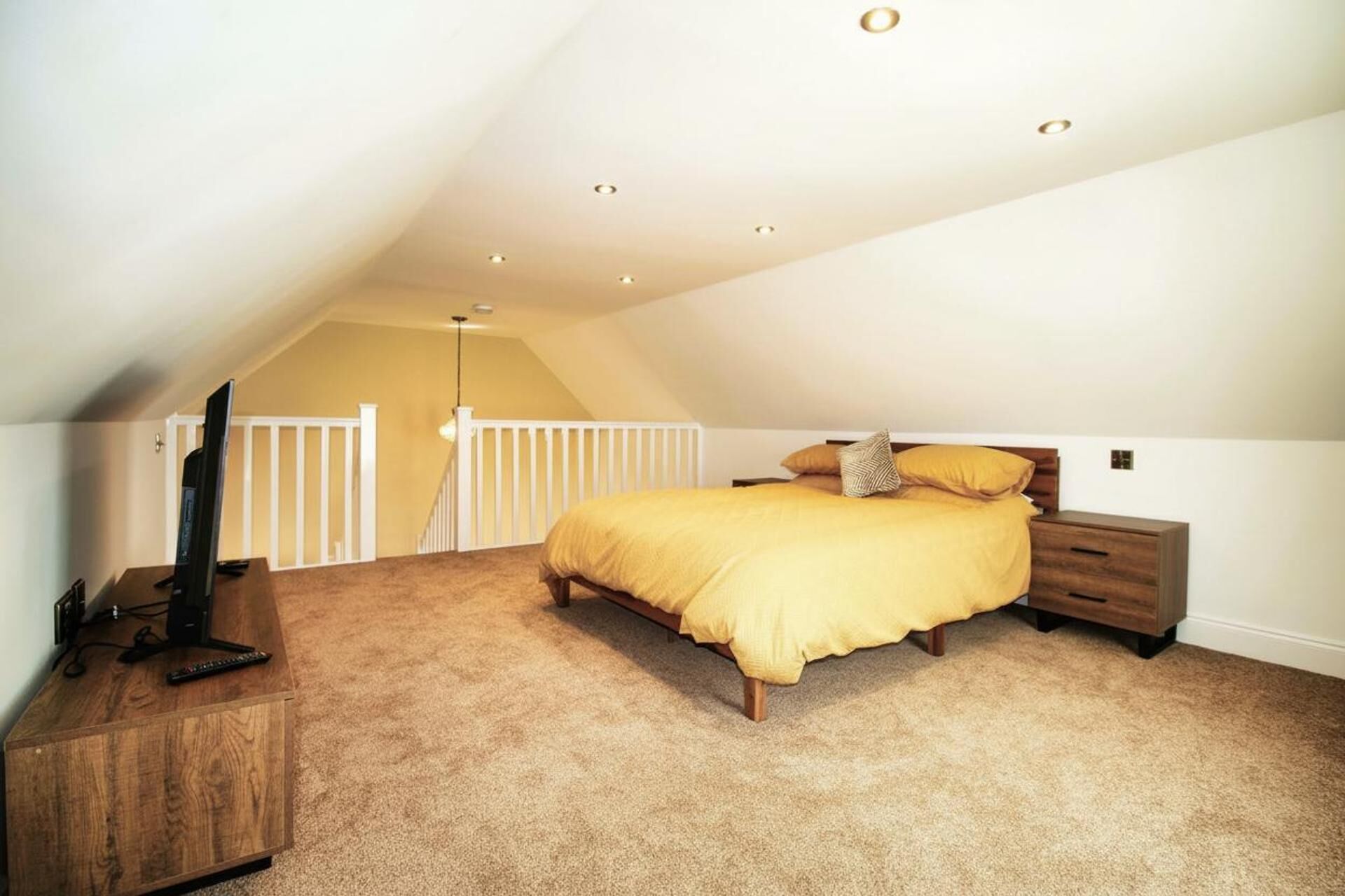 A bedroom with a sloped ceiling, a bed with a yellow bedspread, a wooden nightstand, and a TV on a wooden stand.