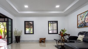 A spacious living room with a black leather sofa, a coffee table, and a painting on the wall. There are two windows with black frames and a wooden stool. - Villa - Sleeps 8 - Terrace - Beach Close (Ko Lanta)
