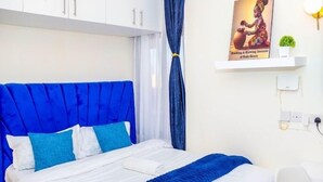 A bedroom with a bed, white and blue bedding, a blue headboard, a white wardrobe, a blue curtain, a wall-mounted shelf with a poster, and a small plant. - Elclassico Studio Jkia Airport View (Nairobi)
