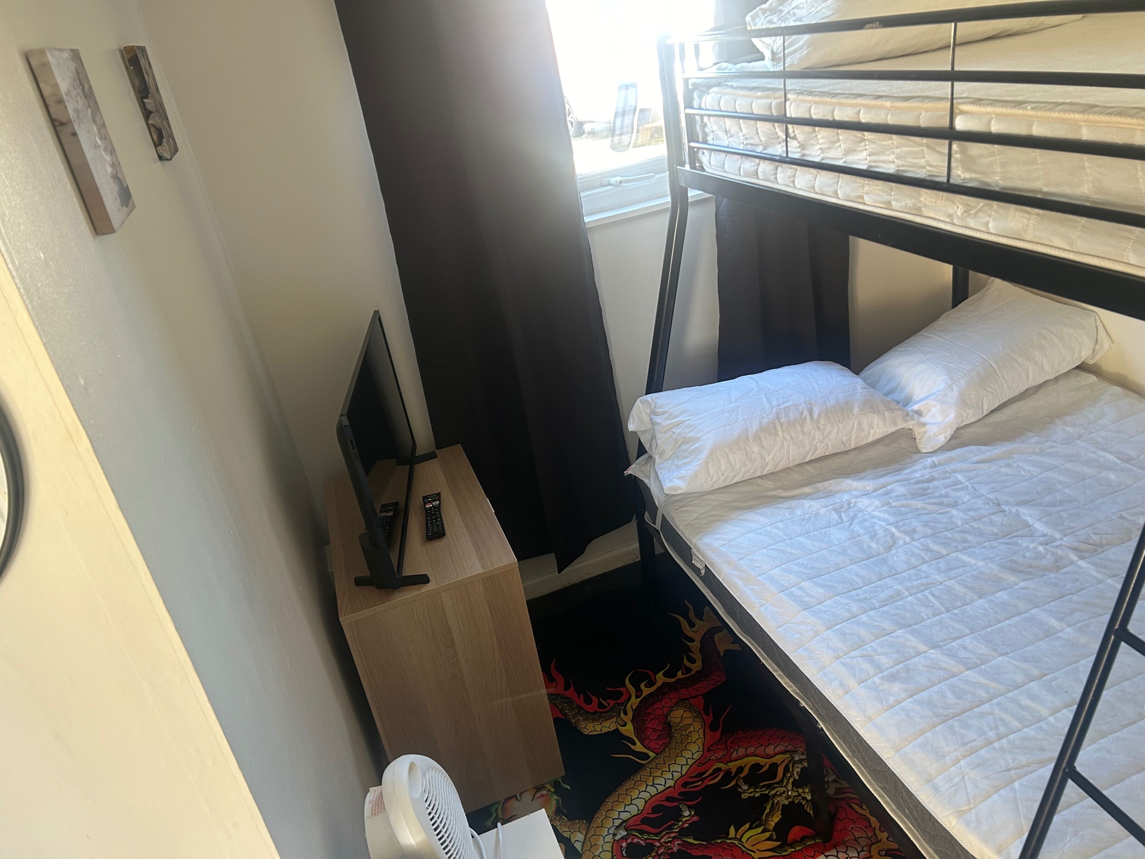 A bunk bed with white bedding, a desk with a computer and remote control, a fan, and a decorative dragon rug.