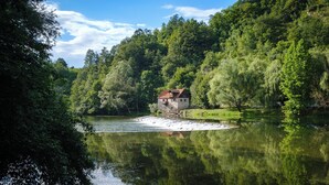 Une maison de campagne en pierre, avec un toit rouge, située au bord d’une rivière paisible, et entourée d’une végétation luxuriante et d’arbres.