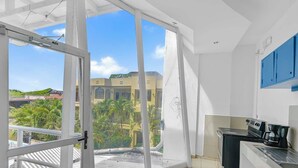 A balcony with a sliding glass door, a kitchen with a stove and coffee maker, and a view of a building with palm trees. - Surfo House (Tamarindo)