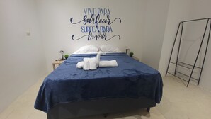 A neatly made bed with a blue bedspread and a swan towel art. There is a wall decoration with text and a small table with a plant and a ring. - Surfo House (Tamarindo)