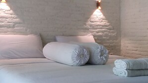 Een net opgemaakt bed met witte lakens en kussens, twee wandlampen en een bakstenen muur.