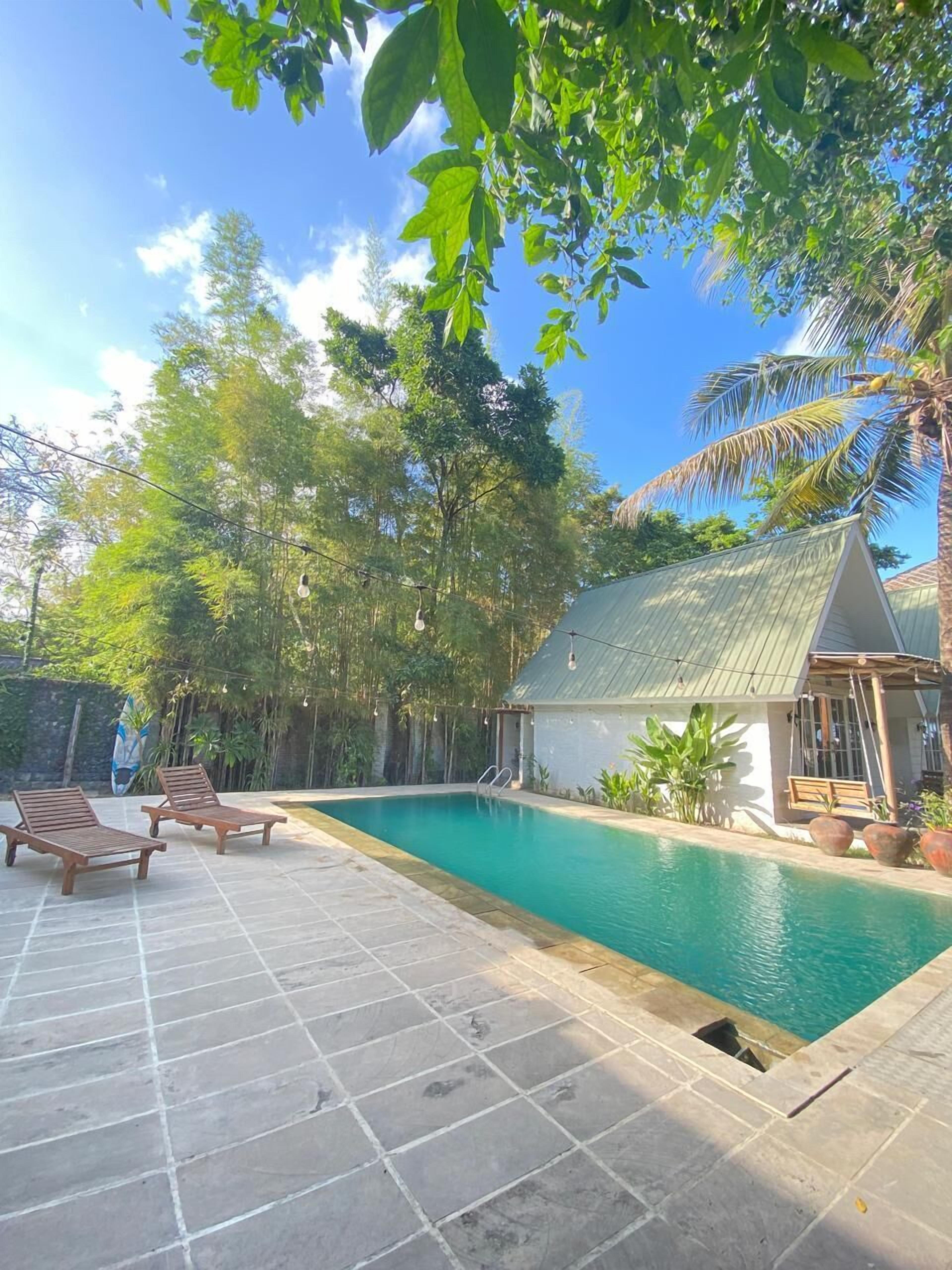 A swimming pool with two wooden loungers, surrounded by lush greenery and a small building with a sloped roof.