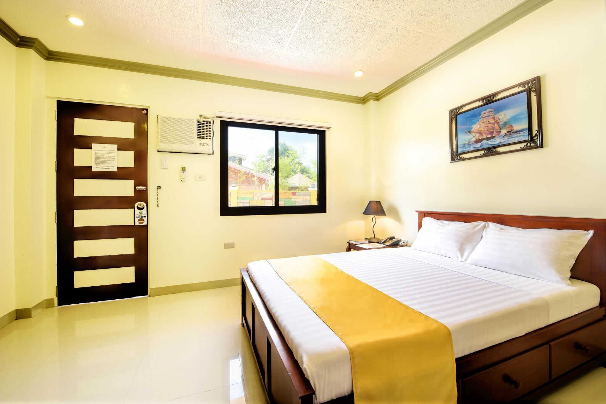 Cama bem arrumada com um cobertor amarelo, cabeceira de madeira, mesa de cabeceira com abajur e janela com vista para prédios.