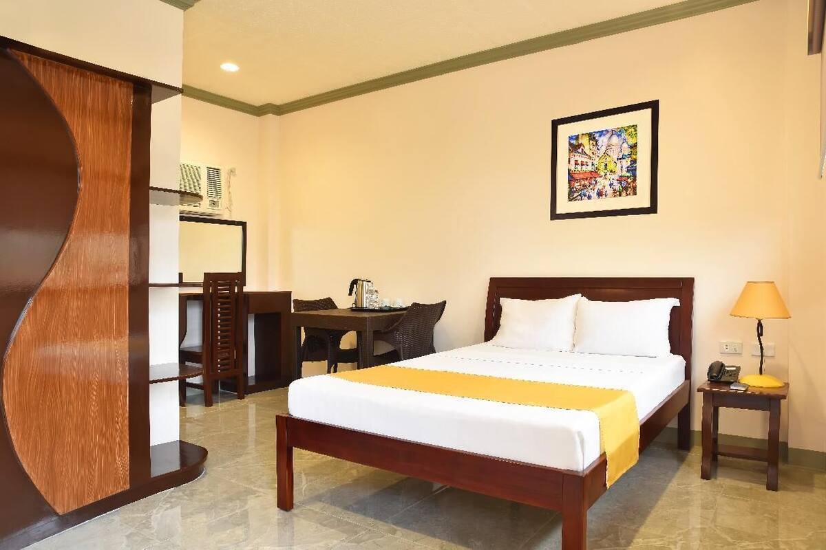 A hotel room with a bed, a desk, a chair, a table, and a framed picture on the wall.