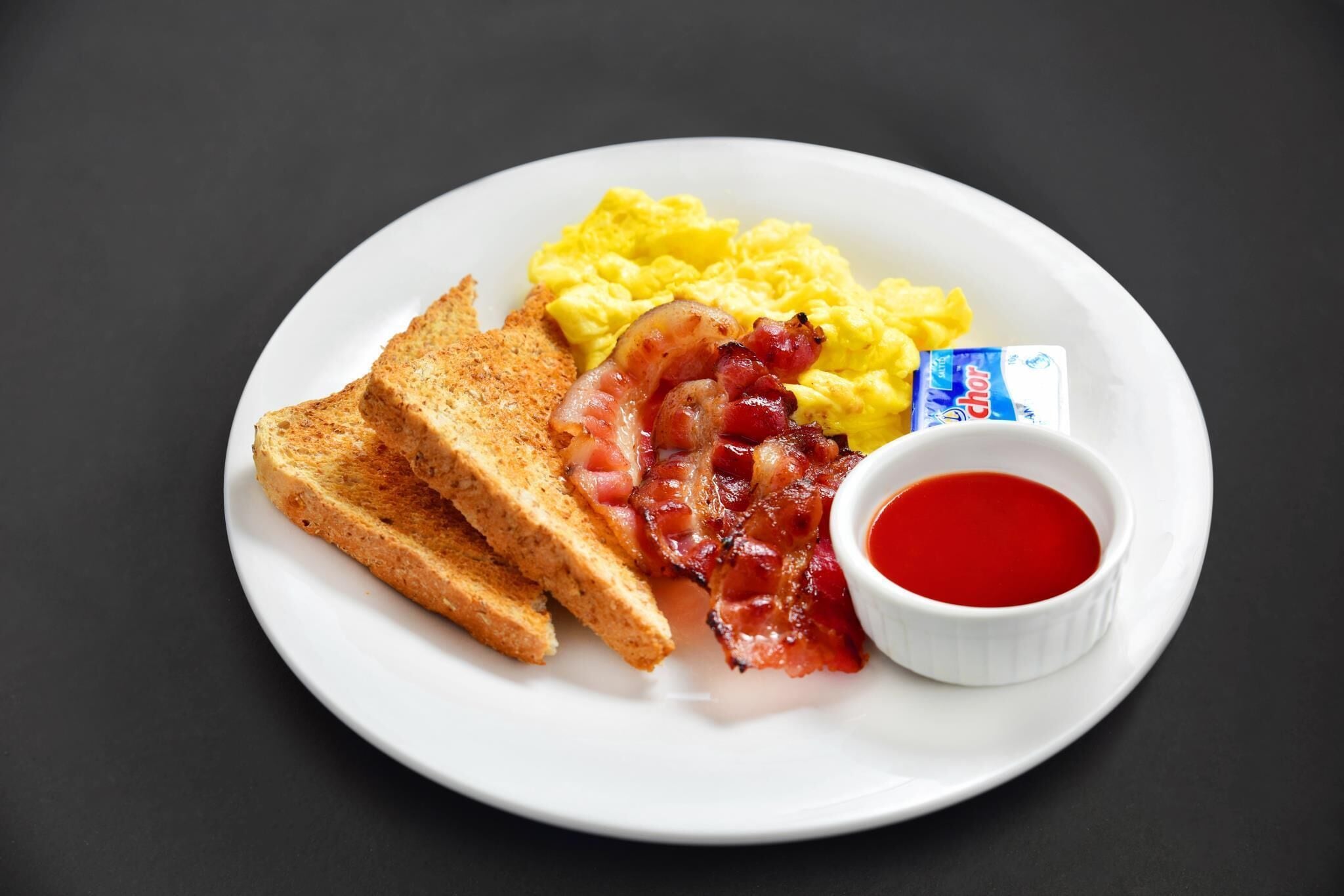 A plate with scrambled eggs, bacon, toast, and a small bowl of jam.