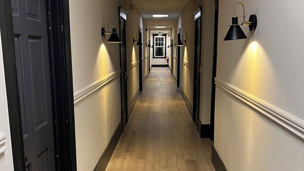 Een lange, smalle hotelgang met houten vloer, deuren met zwarte omlijsting en wandlampen.