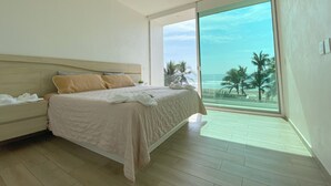 Un dormitorio con una cama grande, una mesita de noche y vistas a la playa a través de una puerta corrediza de vidrio.