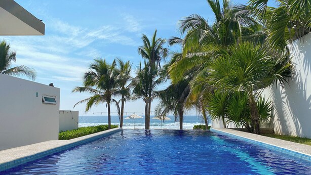 Piscina con vista al océano, rodeada de palmeras y un edificio blanco.