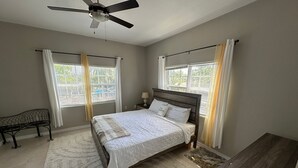 Ein Schlafzimmer mit Bett, Nachttisch, Schreibtisch, Deckenventilator und zwei mit Vorhängen versehenen Fenstern.