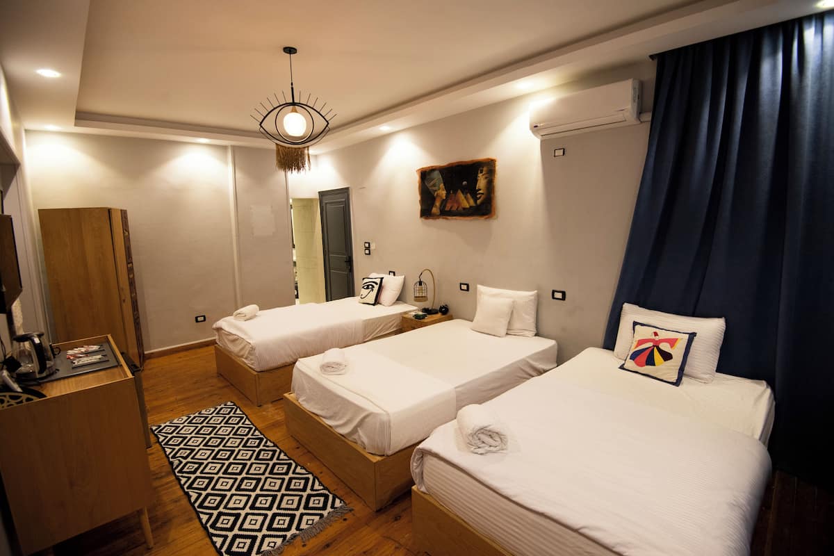 Habitación de hotel con tres camas individuales, un armario de madera, un escritorio con silla y una alfombra estampada.