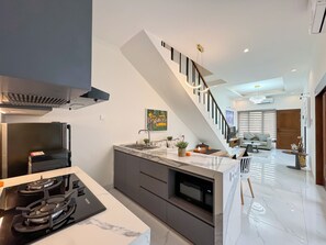 A modern kitchen with a gas stove, marble countertop, and a built-in oven. A staircase leads to a living area with a sofa and a TV. - De Amelia Seminyak Villa by Nagisa Bali (Denpasar)
