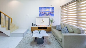 A modern living room with a marble coffee table, a beige sofa with patterned cushions, a staircase with wooden railings, a framed artwork above the sofa, and a ceiling light fixture. - De Amelia Seminyak Villa by Nagisa Bali (Denpasar)