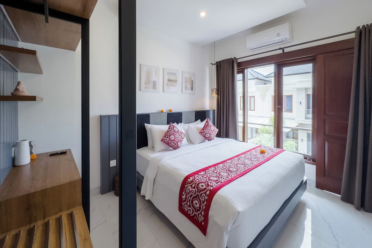 A hotel room with a large bed, a wooden bench, a small table with a vase and a remote control, and an air conditioning unit on the wall.