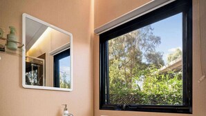 A compact bathroom with a toilet, sink, and mirror. A window provides a view of greenery outside. - Manna Gum Retreat by Tiny Away (Cudgee)