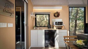 A compact kitchen with a dishwasher, microwave, and a small window overlooking a forest. - Manna Gum Retreat by Tiny Away (Cudgee)