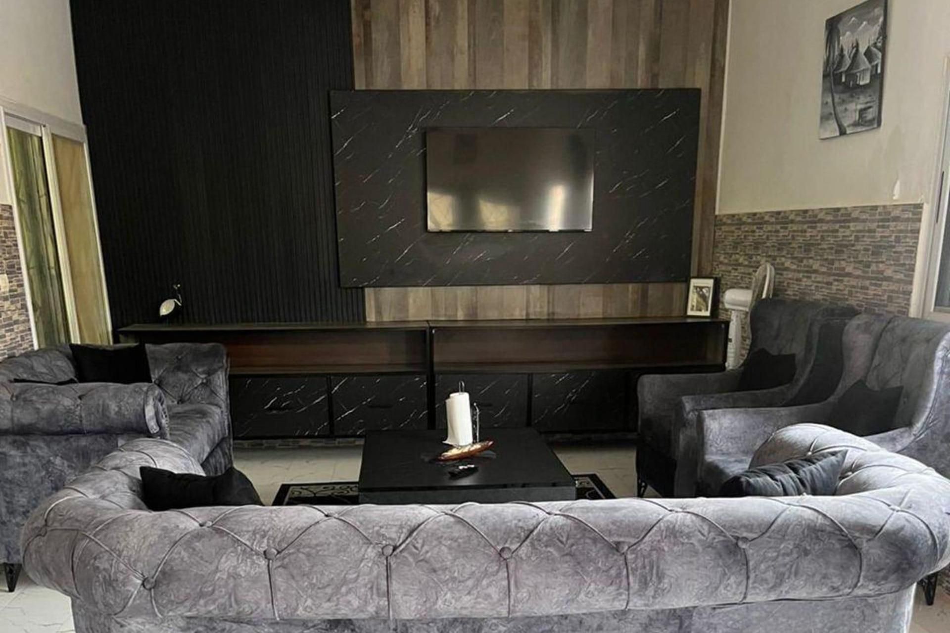 Un salon moderne avec un grand téléviseur à écran plat fixé sur un mur sombre, deux canapés gris rembourrés, une petite table basse noire et une cheminée située sous le téléviseur.