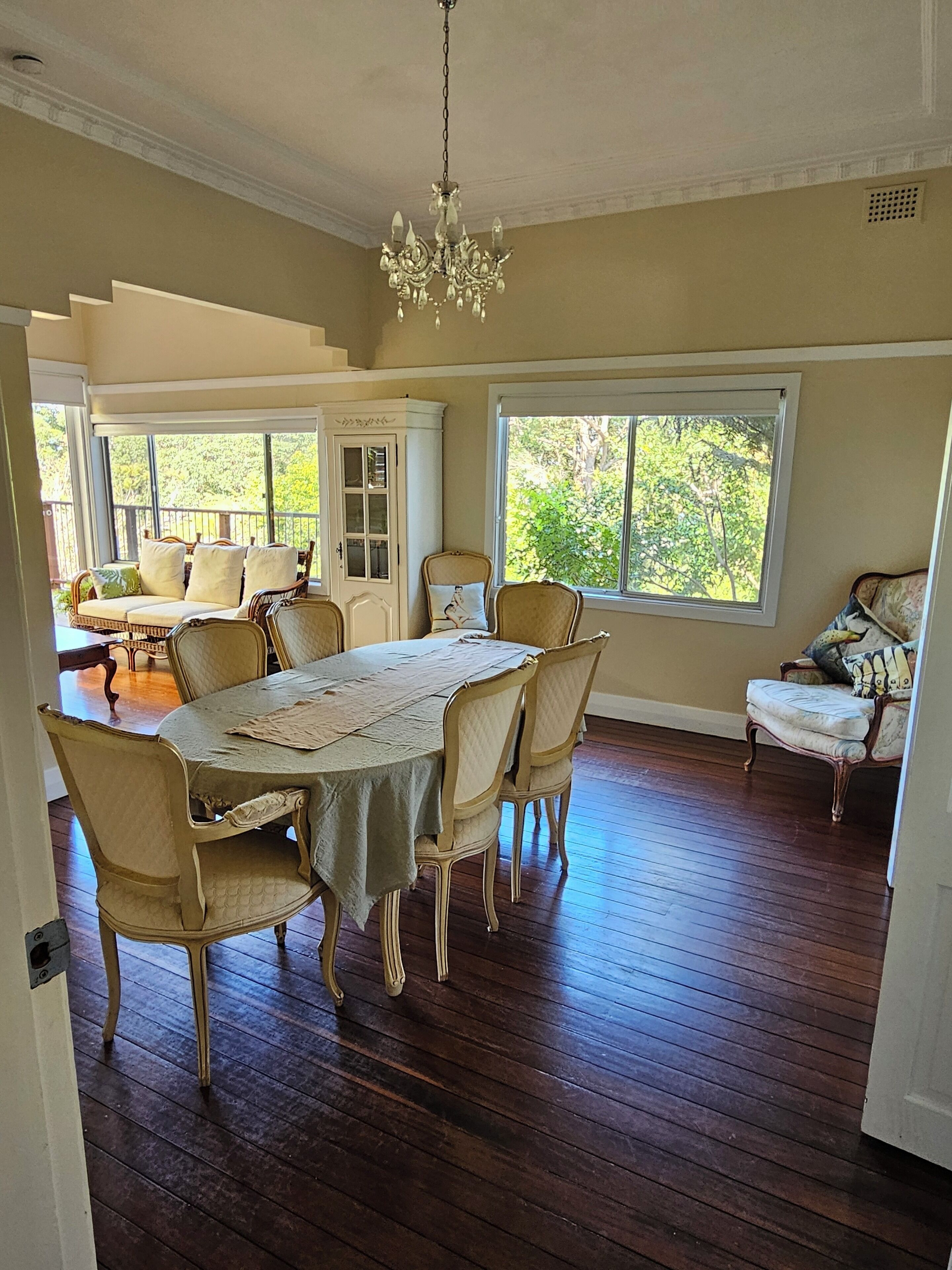 A dining room with a large table, chairs, and a chandelier. There are windows with views of greenery outside.