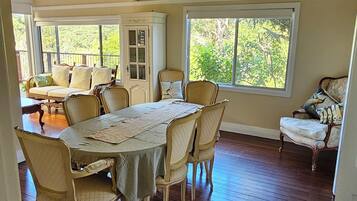 A dining room with a large table, chairs, and a chandelier. There are windows with views of greenery outside.