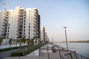 A modern high-rise residential building with balconies, a waterfront promenade, and a clear sky.