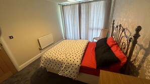 Makuuhuoneessa on sänky, patteri, ikkuna, jossa on verhot, tuoli ja puinen yöpöytä.