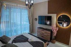 Una habitación de hotel moderna con una cama grande, un televisor de pantalla plana, un aparador de madera, un espejo y un ventanal con cortinas translúcidas.