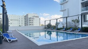 A rooftop swimming pool with blue lounge chairs, a clear sky, and a modern building in the background. - Masdar Oasis 1 B1 - 831 - Jade 2BR Oasis (Abu Dhabi)
