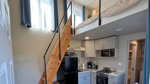 Cocina pequeña con estufa integrada, microondas y refrigerador. Una escalera de madera conduce a un altillo con cama.