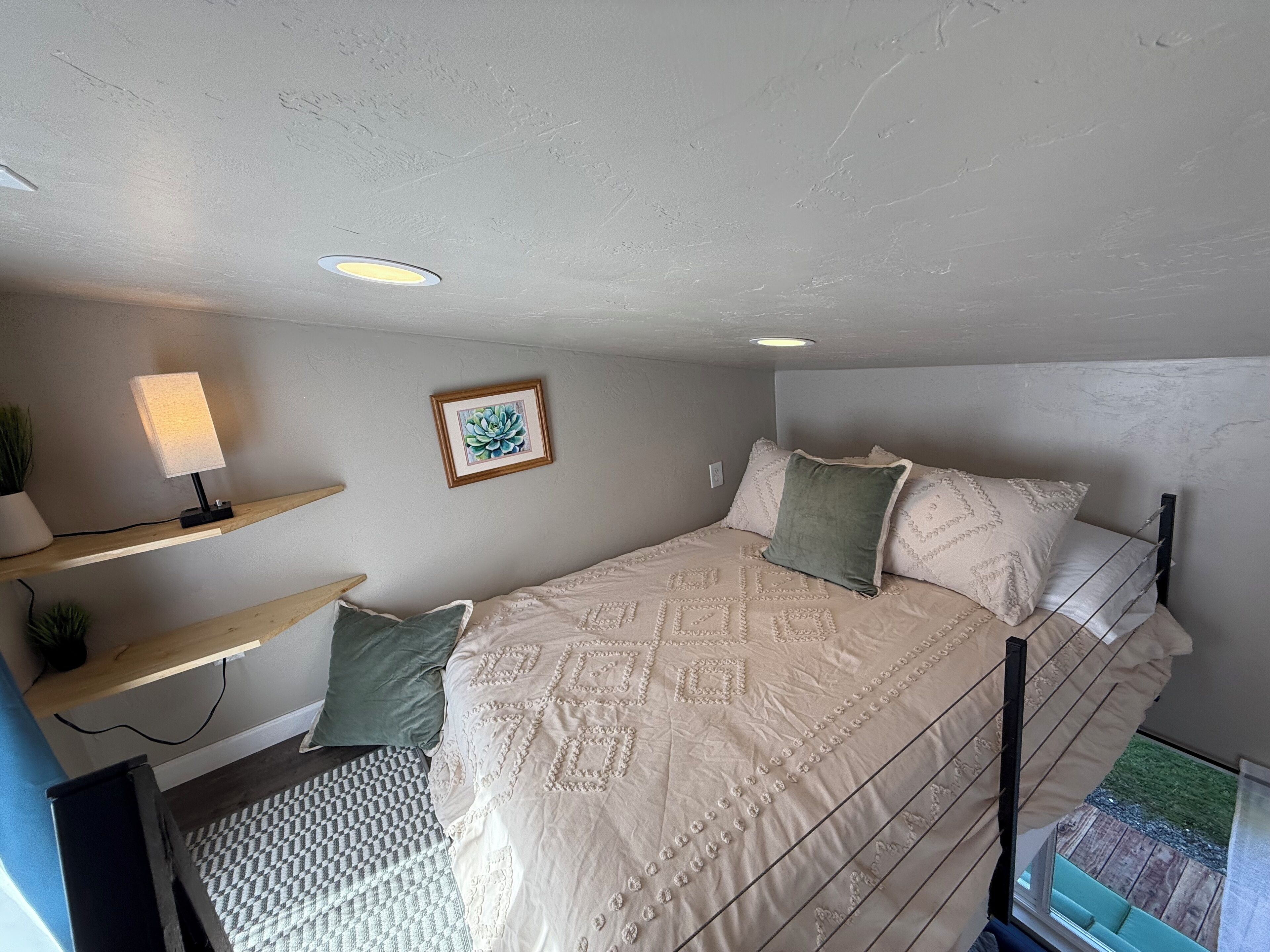 Une chambre à coucher avec un lit, un bureau avec une lampe, un tableau encadré au mur et une fenêtre donnant sur l’extérieur.