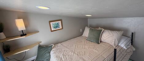 Une chambre à coucher avec un lit, un bureau avec une lampe, un tableau encadré au mur et une fenêtre donnant sur l’extérieur.