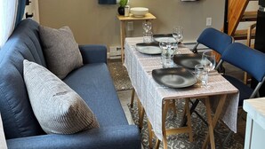 Una sala de estar acogedora con un sofá azul, una mesa de comedor lista para cuatro comensales y un televisor sobre la ventana.