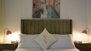 A hotel room with a bed, two bedside tables, a nightstand, a painting of a canal, and a pillow with a crest. - City Nest - 2BR apartment. (Abu Dhabi)