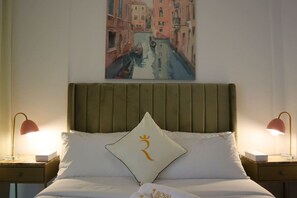 A hotel room with a bed, two bedside tables, a nightstand, a painting of a canal, and a pillow with a crest.
