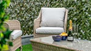 A wicker chair with a cushion, a glass table with a bowl and bottle, and a bottle of champagne.