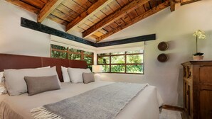 Un dormitorio con una cama grande, vigas de madera en el techo, un ventanal con vistas a la vegetación y un armario de madera.