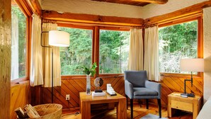 Ein gemütliches Zimmer mit Holzwänden und -böden, einem bequemen Sessel, einem kleinen Tisch mit Lampe und einem Fenster mit Vorhängen.