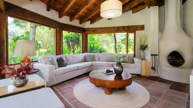 A spacious living room with a wooden ceiling, large windows, a white sofa, a round coffee table, and a fireplace. - Casa Abi - Garden and River Retreat (Valle de Bravo)