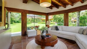 A spacious living room with a white sofa, a round wooden coffee table, and a large window offering a view of a garden. - Casa Abi - Garden and River Retreat (Valle de Bravo)