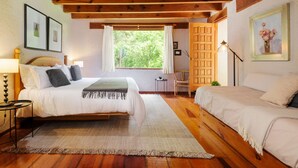 A bedroom with two beds, wooden flooring, a large window with curtains, a door with a grid pattern, and framed pictures on the walls. - Casa Abi - Garden and River Retreat (Valle de Bravo)
