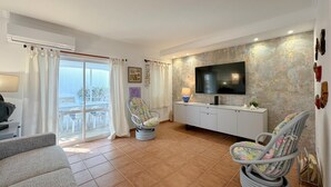 Ein Wohnzimmer mit einer Couch, zwei Sesseln, einem Fernseher und einem Balkon mit Vorhängen.