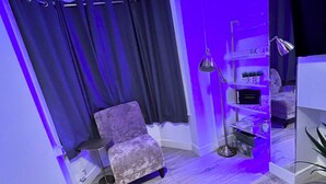 A room with a purple-lit corner, a plush chair, a small table, and a shelf with decorative items. - Gillingham - 3 Beds - Sleeps 6 - Garden - Wifi (Gillingham)