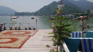 Un muelle de madera con plantas en macetas y un tapete, con vista a un lago con barcos y una montaña al fondo.