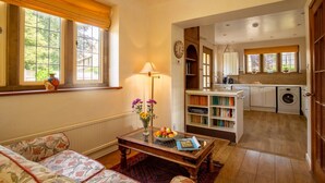 A cozy living room with a sofa, a coffee table with a vase of flowers, and a view into the kitchen area. - Arlington House - sleeps 6 guests  in 3 bedrooms (Lower Slaughter)