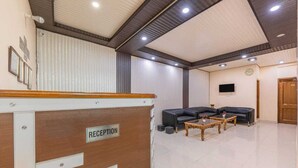 A reception area with a wooden reception desk labeled "RECEPTION," a black leather sofa, two wooden coffee tables, and a television mounted on the wall. - Hotel Charming Hills By Him Haults Hospitality (Shimla)