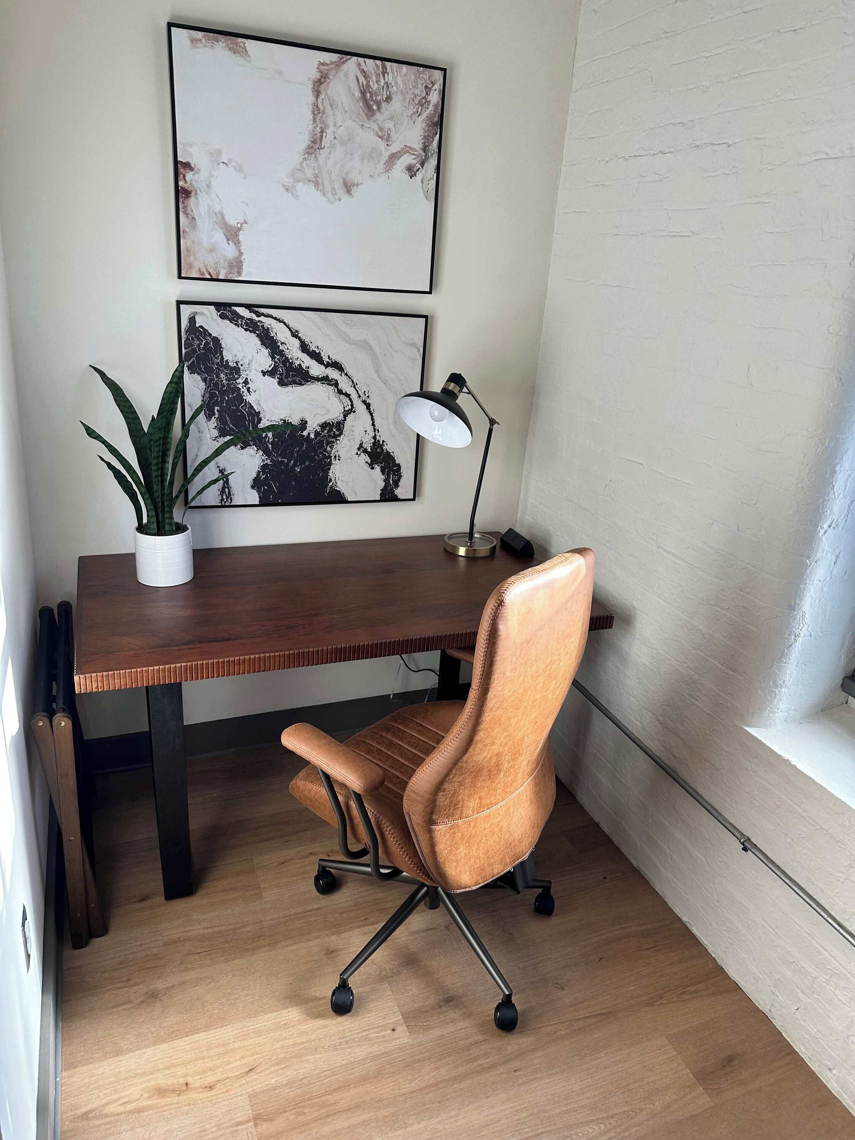 A workspace with a wooden desk, a brown leather chair, a potted plant, and two framed abstract artworks on the wall.